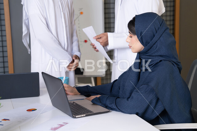 Studying and developing new projects, the work environment in UAE companies, using modern technical equipment, achieving company goals, a meeting and discussion at the company headquarters, an Emirati Gulf Arab businesswoman wearing a hijab and abaya working on a laptop with her colleagues inside the office