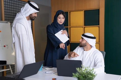 اجتماع ومناقشة عمل في مقر الشركة، رجل أعمال عربي خليجي اماراتي يرتدي الكندورة والغترة يقوم بالعمل على الحاسب المحمول مع زملاءه داخل المكتب، تطوير ودراسة المشاريع الجديدة في شركات الامارات العربية المتحدة، استخدام جهاز تقني حديث، جمع البيانات وحل المشكلات