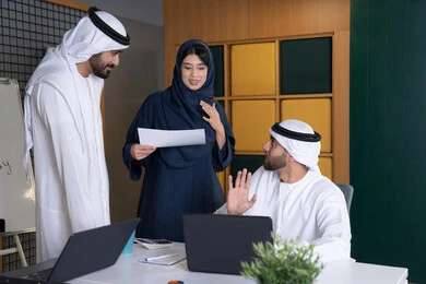 تحليل البيانات وحل المشكلات، اجتماع ومناقشة عمل في مقر الشركة، رجل أعمال عربي خليجي اماراتي يرتدي الكندورة والغترة يقوم بالعمل على الحاسب المحمول مع زملاءه داخل المكتب، تطوير ودراسة المشاريع الجديدة في شركات الامارات العربية المتحدة، استخدام جهاز تقني حديث