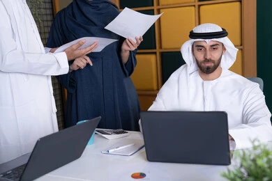 تطوير ودراسة المشاريع الجديدة في شركات الامارات العربية المتحدة، استخدام جهاز تقني حديث، جمع البيانات وحل المشكلات، اجتماع ومناقشة عمل في مقر الشركة، رجل أعمال عربي خليجي اماراتي يرتدي الكندورة والغترة يقوم بالعمل على الحاسب المحمول مع زملاءه داخل المكتب