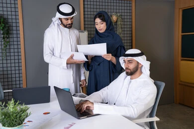 A business meeting and discussion at the company headquarters. An Emirati Gulf Arab businessman wearing a kandura and ghutra is working on a laptop with his colleagues inside the office. Developing and studying new projects in UAE companies, using a modern technical device, collecting data and solving problems.