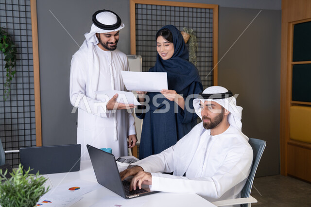 تحليل البيانات وحل المشكلات، اجتماع ومناقشة عمل في مقر الشركة، رجل أعمال عربي خليجي اماراتي يرتدي الكندورة والغترة يقوم بالعمل على الحاسب المحمول مع زملاءه داخل المكتب، تطوير ودراسة المشاريع الجديدة في شركات الامارات العربية المتحدة، استخدام جهاز تقني حديث