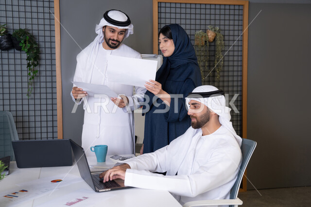 جمع البيانات وحل المشكلات، اجتماع ومناقشة عمل في مقر الشركة، رجل أعمال عربي خليجي اماراتي يرتدي الكندورة والغترة يقوم بالعمل على الحاسب المحمول مع زملاءه داخل المكتب، تطوير ودراسة المشاريع الجديدة في شركات الامارات العربية المتحدة، استخدام جهاز تقني حديث