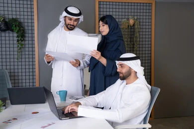 جمع البيانات وحل المشكلات، اجتماع ومناقشة عمل في مقر الشركة، رجل أعمال عربي خليجي اماراتي يرتدي الكندورة والغترة يقوم بالعمل على الحاسب المحمول مع زملاءه داخل المكتب، تطوير ودراسة المشاريع الجديدة في شركات الامارات العربية المتحدة، استخدام جهاز تقني حديث