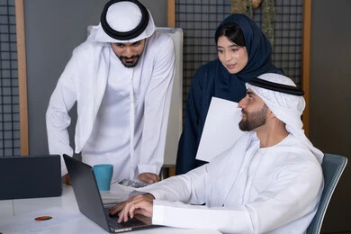 استخدام جهاز تقني حديث، جمع البيانات وحل المشكلات، اجتماع ومناقشة عمل في مقر الشركة، رجل أعمال عربي خليجي اماراتي يرتدي الكندورة والغترة يقوم بالعمل على الحاسب المحمول مع زملاءه داخل المكتب، تطوير ودراسة المشاريع الجديدة في شركات الامارات العربية المتحدة