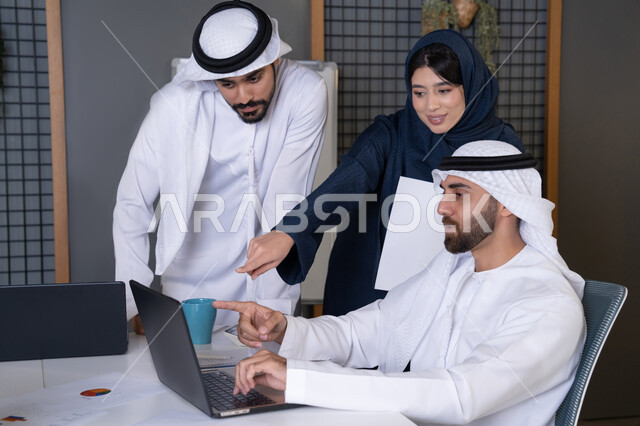 الاشارة الى الحاسب المحمول، استخدام جهاز تقني حديث، جمع البيانات وحل المشكلات، اجتماع ومناقشة عمل في مقر الشركة، رجل أعمال عربي خليجي اماراتي يرتدي الكندورة والغترة يقوم بالتحدث مع زملاءه داخل المكتب، تطوير ودراسة المشاريع الجديدة في شركات الامارات العربية المتحدة