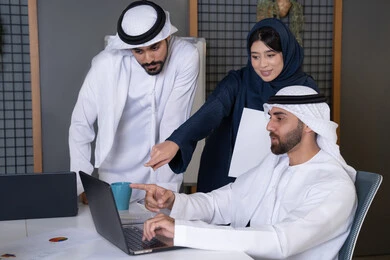 الاشارة الى الحاسب المحمول، استخدام جهاز تقني حديث، جمع البيانات وحل المشكلات، اجتماع ومناقشة عمل في مقر الشركة، رجل أعمال عربي خليجي اماراتي يرتدي الكندورة والغترة يقوم بالتحدث مع زملاءه داخل المكتب، تطوير ودراسة المشاريع الجديدة في شركات الامارات العربية المتحدة