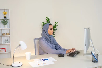 الكتابة على لوحة المفاتيح، مهنة ووظيفة نسائية،  إمرأة عربية خليجية اماراتية محجبة ترتدي عباءة ملونة تجلس خلف الطاولة، سيدة سعودية تستخدم الحاسوب في المكتب، استخدام جهاز تقني حديث ومتطور، ايماءات التركيز والاندماج، ادارة الأعمال وانجاز المهام المطلوبة