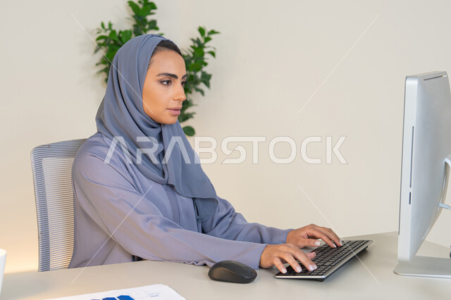 Business administration and completing required tasks, typing on a keyboard, a female profession and job, a veiled Emirati Gulf Arab woman wearing a colorful abaya sitting behind a table, a Saudi woman using a computer in the office, using a modern and advanced technical device, gestures of concentration and integration