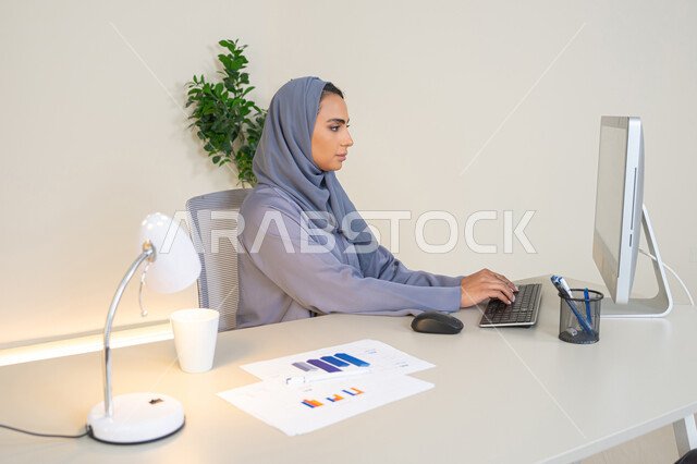 Analysis of statistical data results, a veiled Emirati Gulf Arab woman wearing an abaya sitting behind a table with a group of charts next to her, a Saudi woman taking notes on paper, working on a computer to complete tasks in the office, an office profession and job in Gulf companies, using an advanced technical device