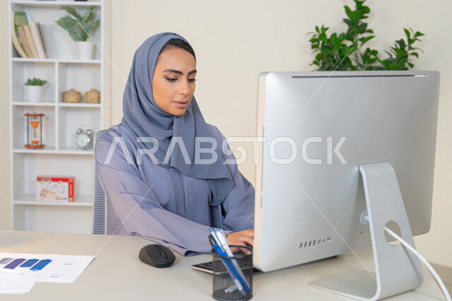 Business administration and completing required tasks, typing on a keyboard, a female profession and job, a veiled Emirati Gulf Arab woman wearing a colorful abaya sitting behind a table, a Saudi woman using a computer in the office, using a modern and advanced technical device, gestures of concentration and integration