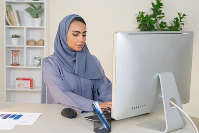 ادارة الأعمال وانجاز المهام المطلوبة، الكتابة على لوحة المفاتيح، مهنة ووظيفة نسائية،  إمرأة عربية خليجية اماراتية محجبة ترتدي عباءة ملونة تجلس خلف الطاولة، سيدة سعودية تستخدم الحاسوب في المكتب، استخدام جهاز تقني حديث ومتطور، ايماءات التركيز والاندماج