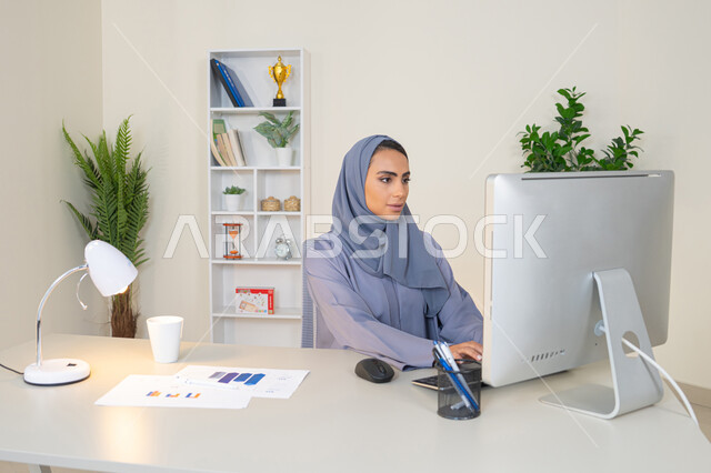 الكتابة على لوحة المفاتيح، مهنة ووظيفة نسائية،  إمرأة عربية خليجية اماراتية محجبة ترتدي عباءة ملونة تجلس خلف الطاولة، سيدة سعودية تستخدم الحاسوب في المكتب، استخدام جهاز تقني حديث ومتطور، ايماءات التركيز والاندماج، ادارة الأعمال وانجاز المهام المطلوبة