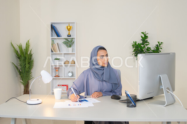 تحليل نتائج البيانات الاحصائية، امراة عربية خليجية اماراتية محجبة ترتدي العباءة تجلس خلف الطاولة وبجانبها مجموعة مخططات، سيدة سعودية تدون الملاحظات على الورق، العمل على الحاسب لانجاز المهام في المكتب، مهنة ووظيفة مكتبية في شركات الخليج، استخدام جهاز تقني متطور