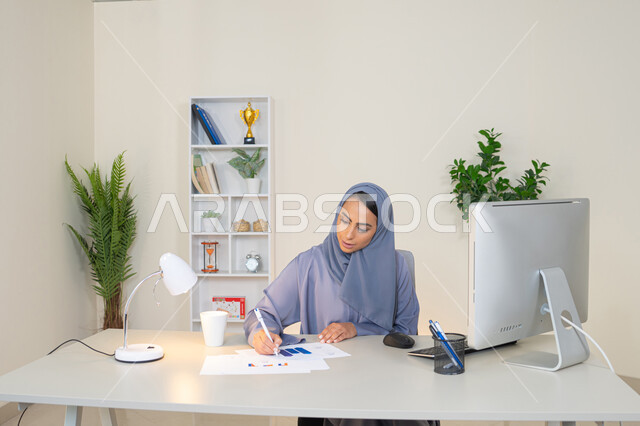 العمل على الحاسب لانجاز المهام في المكتب، وظيفة مكتبية في شركات الخليج العربي، استخدام جهاز تقني حديث ومتطور، تحليل نتائج البيانات الاحصائية، امراة عربية خليجية اماراتية محجبة ترتدي العباءة تجلس خلف الطاولة وبجانبها مجموعة مخططات، سيدة سعودية تدون الملاحظات على الورق