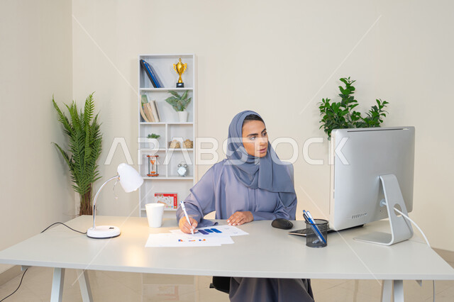 تحليل نتائج البيانات الاحصائية، امراة عربية خليجية اماراتية محجبة ترتدي العباءة تجلس خلف الطاولة وبجانبها مجموعة مخططات، سيدة سعودية تدون الملاحظات على الورق، العمل على الحاسب لانجاز المهام في المكتب، مهنة ووظيفة مكتبية في شركات الخليج، استخدام جهاز تقني متطور