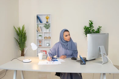 Analysis of statistical data results, a veiled Emirati Gulf Arab woman wearing an abaya sitting behind a table with a group of charts next to her, a Saudi woman taking notes on paper, working on a computer to complete tasks in the office, an office profession and job in Gulf companies, using an advanced technical device