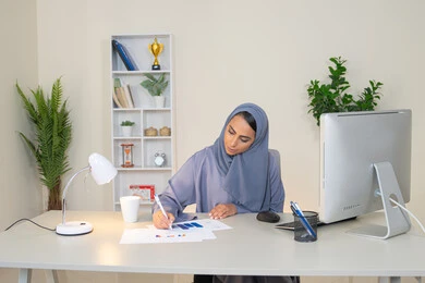 العمل على الحاسب لانجاز المهام في المكتب، سيدة سعودية  تستخدام الالة الحاسبة لاجراء العمليات الحسابية، وظيفة مكتبية في شركات الخليج العربي، تحليل نتائج البيانات الاحصائية، امراة عربية خليجية اماراتية محجبة ترتدي العباءة تجلس خلف الطاولة تدون الملاحظات على الورق