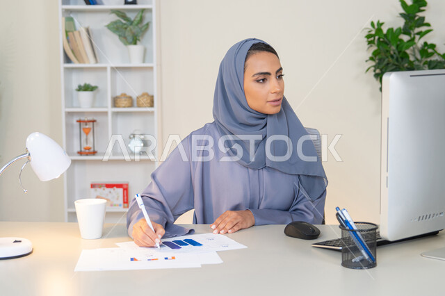 استخدام جهاز تقني متطور، تحليل نتائج البيانات الاحصائية، امراة عربية خليجية اماراتية محجبة ترتدي العباءة تجلس خلف الطاولة وبجانبها مجموعة مخططات، سيدة سعودية تدون الملاحظات على الورق، العمل على الحاسب لانجاز المهام في المكتب، وظيفة مكتبية في شركات الخليج العربي