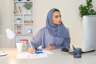 استخدام جهاز تقني متطور، تحليل نتائج البيانات الاحصائية، امراة عربية خليجية اماراتية محجبة ترتدي العباءة تجلس خلف الطاولة وبجانبها مجموعة مخططات، سيدة سعودية تدون الملاحظات على الورق، العمل على الحاسب لانجاز المهام في المكتب، وظيفة مكتبية في شركات الخليج العربي