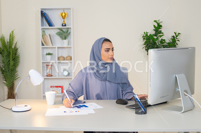 العمل على الحاسب لانجاز المهام في المكتب، وظيفة مكتبية في شركات الخليج العربي، استخدام جهاز تقني حديث ومتطور، تحليل نتائج البيانات الاحصائية، امراة عربية خليجية اماراتية محجبة ترتدي العباءة تجلس خلف الطاولة وبجانبها مجموعة مخططات، سيدة سعودية تدون الملاحظات على الورق