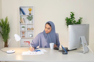 تحليل نتائج البيانات الاحصائية، امراة عربية خليجية اماراتية محجبة ترتدي العباءة تجلس خلف الطاولة وبجانبها مجموعة مخططات، سيدة سعودية تدون الملاحظات على الورق، العمل على الحاسب لانجاز المهام في المكتب، مهنة ووظيفة مكتبية في شركات الخليج، استخدام جهاز تقني متطور