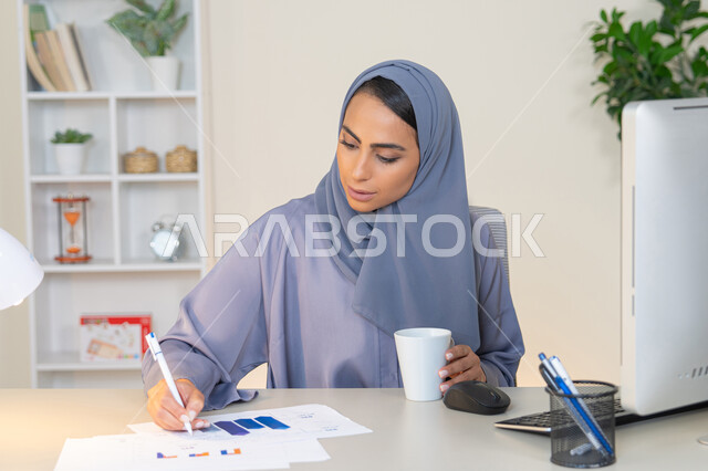 استخدام جهاز تقني متطور، تحليل نتائج البيانات الاحصائية، امراة عربية خليجية اماراتية محجبة ترتدي العباءة تجلس خلف الطاولة وبجانبها مجموعة مخططات، سيدة سعودية تدون الملاحظات على الورق، العمل على الحاسب لانجاز المهام في المكتب، وظيفة مكتبية في شركات الخليج العربي