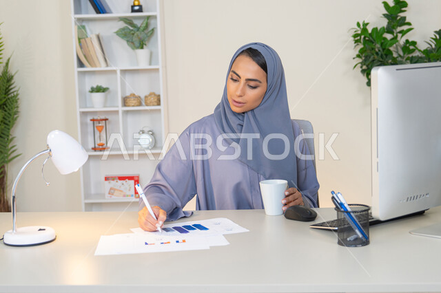 تحليل نتائج البيانات الاحصائية، امراة عربية خليجية اماراتية محجبة ترتدي العباءة تجلس خلف الطاولة وبجانبها مجموعة مخططات، سيدة سعودية تدون الملاحظات على الورق، العمل على الحاسب لانجاز المهام في المكتب، مهنة ووظيفة مكتبية في شركات الخليج، استخدام جهاز تقني متطور