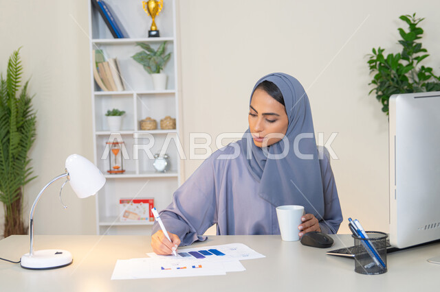 استخدام جهاز تقني متطور، تحليل نتائج البيانات الاحصائية، امراة عربية خليجية اماراتية محجبة ترتدي العباءة تجلس خلف الطاولة وبجانبها مجموعة مخططات، سيدة سعودية تدون الملاحظات على الورق، العمل على الحاسب لانجاز المهام في المكتب، وظيفة مكتبية في شركات الخليج العربي