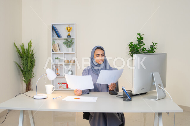 العمل على الحاسب لانجاز المهام في المكتب، وظيفة مكتبية في شركات الخليج العربي، استخدام جهاز تقني حديث ومتطور، تحليل نتائج البيانات الاحصائية، امراة عربية خليجية اماراتية محجبة ترتدي العباءة تجلس خلف الطاولة وبجانبها مجموعة مخططات، سيدة سعودية تدون الملاحظات على الورق