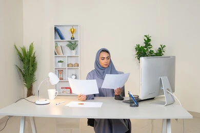 العمل على الحاسب لانجاز المهام في المكتب، وظيفة مكتبية في شركات الخليج العربي، استخدام جهاز تقني حديث ومتطور، تحليل نتائج البيانات الاحصائية، امراة عربية خليجية اماراتية محجبة ترتدي العباءة تجلس خلف الطاولة وبجانبها مجموعة مخططات، سيدة سعودية تدون الملاحظات على الورق