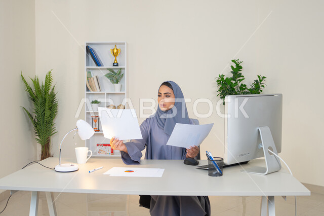 انجاز المهام في المكتب، وظيفة مكتبية في شركات الخليج العربي، استخدام جهاز تقني حديث ومتطور، تحليل نتائج البيانات الاحصائية، امراة عربية خليجية اماراتية محجبة ترتدي العباءة تجلس خلف الطاولة وبجانبها مجموعة مخططات تعمل على الحاسب، سيدة سعودية تدون الملاحظات على الورق