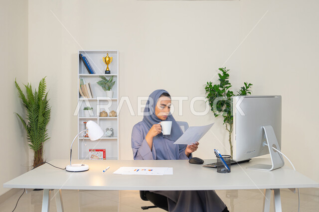 استخدام جهاز تقني حديث ومتطور، تحليل نتائج البيانات الاحصائية، امراة عربية خليجية اماراتية محجبة ترتدي العباءة تجلس خلف الطاولة وبجانبها مجموعة مخططات تعمل على الحاسب لانجاز المهام في المكتب، سيدة سعودية تدون الملاحظات على الورق، وظيفة مكتبية في شركات الخليج العربي