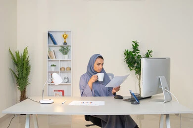 Using a modern and advanced technical device, analyzing the results of statistical data, a veiled Emirati Gulf Arab woman wearing an abaya sitting behind a table and next to her a group of computer-generated charts to complete tasks in the office, a Saudi woman taking notes on paper, an office job in a Gulf Arab company