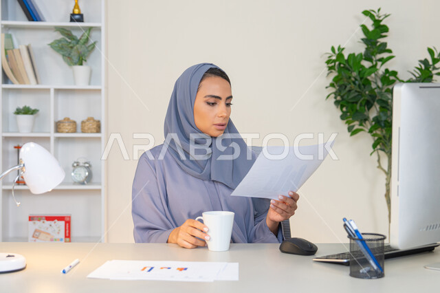 وظيفة مكتبية في شركات الخليج العربي، استخدام جهاز تقني حديث ومتطور، تحليل نتائج البيانات الاحصائية، امراة عربية خليجية اماراتية محجبة ترتدي العباءة تجلس خلف الطاولة وبجانبها مجموعة مخططات تعمل على الحاسب لانجاز المهام في المكتب، سيدة سعودية تدون الملاحظات على الورق