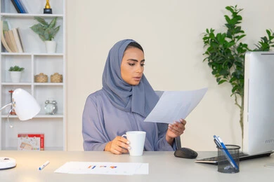 Office job in a Gulf company, using a modern and advanced technical device, analyzing statistical data results, a veiled Emirati Gulf Arab woman wearing an abaya sitting behind a table and next to her a group of computer charts to complete tasks in the office, a Saudi woman taking notes on paper