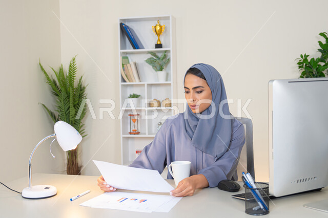 انجاز المهام في المكتب، وظيفة مكتبية في شركات الخليج العربي، استخدام جهاز تقني حديث ومتطور، تحليل نتائج البيانات الاحصائية، امراة عربية خليجية اماراتية محجبة ترتدي العباءة تجلس خلف الطاولة وبجانبها مجموعة مخططات تعمل على الحاسب، سيدة سعودية تدون الملاحظات على الورق