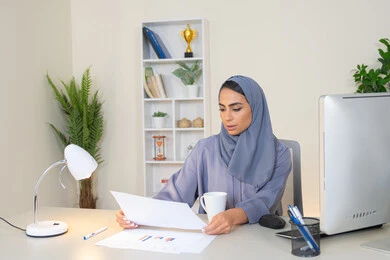 انجاز المهام في المكتب، وظيفة مكتبية في شركات الخليج العربي، استخدام جهاز تقني حديث ومتطور، تحليل نتائج البيانات الاحصائية، امراة عربية خليجية اماراتية محجبة ترتدي العباءة تجلس خلف الطاولة وبجانبها مجموعة مخططات تعمل على الحاسب، سيدة سعودية تدون الملاحظات على الورق