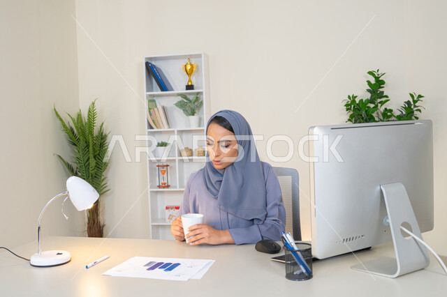 العمل على الحاسب لانجاز المهام في المكتب، وظيفة مكتبية في شركات الخليج العربي، استخدام جهاز تقني حديث ومتطور، تحليل نتائج البيانات الاحصائية، امراة عربية خليجية اماراتية محجبة ترتدي العباءة تجلس خلف الطاولة وبجانبها مجموعة مخططات، سيدة سعودية تدون الملاحظات على الورق