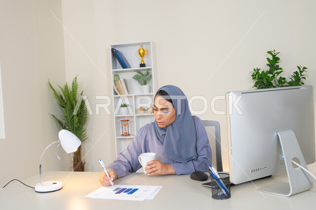 استخدام جهاز تقني حديث ومتطور، تحليل نتائج البيانات الاحصائية، امراة عربية خليجية اماراتية محجبة ترتدي العباءة تجلس خلف الطاولة وبجانبها مجموعة مخططات تعمل على الحاسب لانجاز المهام في المكتب، سيدة سعودية تدون الملاحظات على الورق، وظيفة مكتبية في شركات الخليج العربي