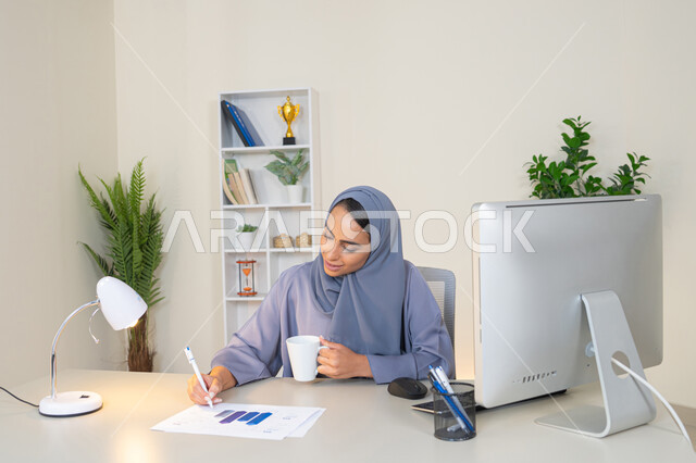 انجاز المهام في المكتب، وظيفة مكتبية في شركات الخليج العربي، استخدام جهاز تقني حديث ومتطور، تحليل نتائج البيانات الاحصائية، امراة عربية خليجية اماراتية محجبة ترتدي العباءة تجلس خلف الطاولة وبجانبها مجموعة مخططات تعمل على الحاسب، سيدة سعودية تدون الملاحظات على الورق