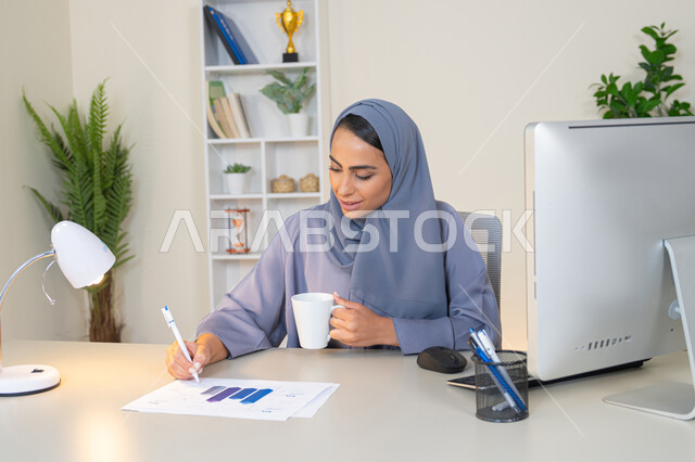 استخدام جهاز تقني حديث ومتطور، تحليل نتائج البيانات الاحصائية، امراة عربية خليجية اماراتية محجبة ترتدي العباءة تجلس خلف الطاولة وبجانبها مجموعة مخططات تعمل على الحاسب لانجاز المهام في المكتب، سيدة سعودية تدون الملاحظات على الورق، وظيفة مكتبية في شركات الخليج العربي