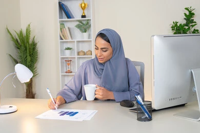 استخدام جهاز تقني حديث ومتطور، تحليل نتائج البيانات الاحصائية، امراة عربية خليجية اماراتية محجبة ترتدي العباءة تجلس خلف الطاولة وبجانبها مجموعة مخططات تعمل على الحاسب لانجاز المهام في المكتب، سيدة سعودية تدون الملاحظات على الورق، وظيفة مكتبية في شركات الخليج العربي
