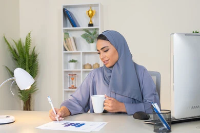 وظيفة مكتبية في شركات الخليج العربي، استخدام جهاز تقني حديث ومتطور، تحليل نتائج البيانات الاحصائية، امراة عربية خليجية اماراتية محجبة ترتدي العباءة تجلس خلف الطاولة وبجانبها مجموعة مخططات تعمل على الحاسب لانجاز المهام في المكتب، سيدة سعودية تدون الملاحظات على الورق