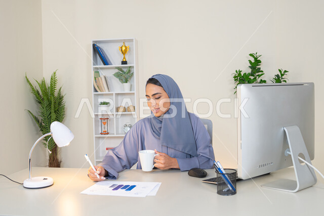 انجاز المهام في المكتب، وظيفة مكتبية في شركات الخليج العربي، استخدام جهاز تقني حديث ومتطور، تحليل نتائج البيانات الاحصائية، امراة عربية خليجية اماراتية محجبة ترتدي العباءة تجلس خلف الطاولة وبجانبها مجموعة مخططات تعمل على الحاسب، سيدة سعودية تدون الملاحظات على الورق
