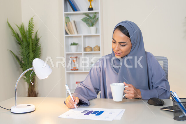 وظيفة مكتبية في شركات الخليج العربي، استخدام جهاز تقني حديث ومتطور، تحليل نتائج البيانات الاحصائية، امراة عربية خليجية اماراتية محجبة ترتدي العباءة تجلس خلف الطاولة وبجانبها مجموعة مخططات تعمل على الحاسب لانجاز المهام في المكتب، سيدة سعودية تدون الملاحظات على الورق