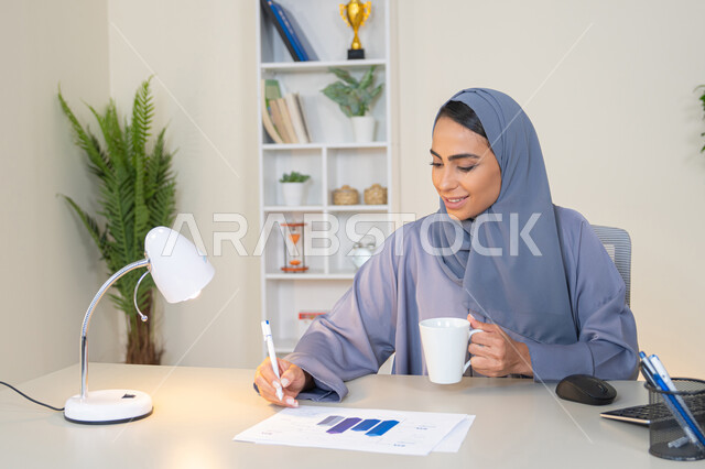 استخدام جهاز تقني حديث ومتطور، تحليل نتائج البيانات الاحصائية، امراة عربية خليجية اماراتية محجبة ترتدي العباءة تجلس خلف الطاولة وبجانبها مجموعة مخططات تعمل على الحاسب لانجاز المهام في المكتب، سيدة سعودية تدون الملاحظات على الورق، وظيفة مكتبية في شركات الخليج العربي