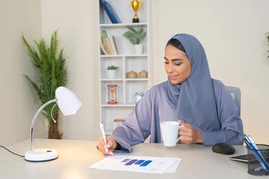استخدام جهاز تقني حديث ومتطور، تحليل نتائج البيانات الاحصائية، امراة عربية خليجية اماراتية محجبة ترتدي العباءة تجلس خلف الطاولة وبجانبها مجموعة مخططات تعمل على الحاسب لانجاز المهام في المكتب، سيدة سعودية تدون الملاحظات على الورق، وظيفة مكتبية في شركات الخليج العربي