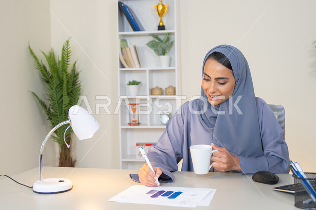 وظيفة مكتبية في شركات الخليج العربي، استخدام جهاز تقني حديث ومتطور، تحليل نتائج البيانات الاحصائية، امراة عربية خليجية اماراتية محجبة ترتدي العباءة تجلس خلف الطاولة وبجانبها مجموعة مخططات تعمل على الحاسب لانجاز المهام في المكتب، سيدة سعودية تدون الملاحظات على الورق