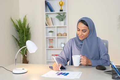 وظيفة مكتبية في شركات الخليج العربي، استخدام جهاز تقني حديث ومتطور، تحليل نتائج البيانات الاحصائية، امراة عربية خليجية اماراتية محجبة ترتدي العباءة تجلس خلف الطاولة وبجانبها مجموعة مخططات تعمل على الحاسب لانجاز المهام في المكتب، سيدة سعودية تدون الملاحظات على الورق